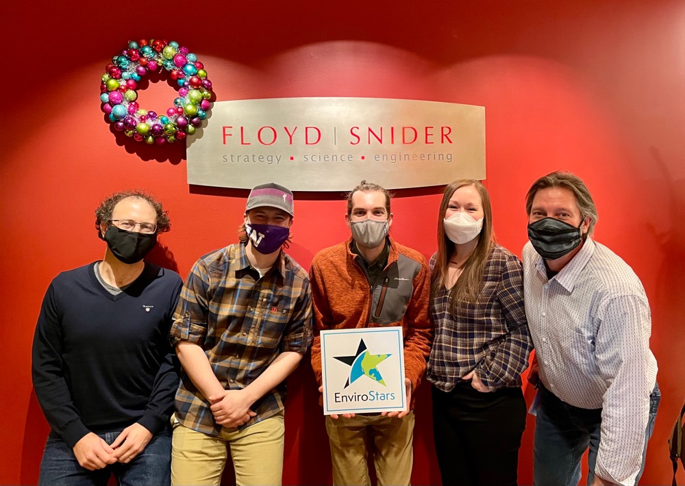 Five members of the Green Team stand in front of a red wall with the Floyd|Snider logo on a metal sign above their heads. Each wears casual clothing and a mask. An EnviroStars sign is being held by the person in the middle.